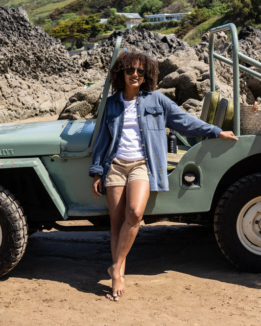 Wearing shorts, a t-shirt, and the Saltrock Maddie Women's Oversized Shirt in Blue, a person stands beside a vintage jeep on a rocky beach.