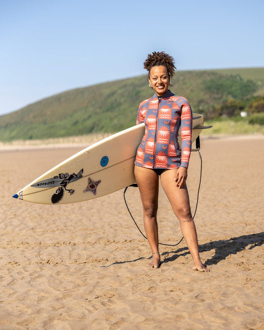 A woman wearing the Saltrock Equinox Womens Wetsuit Jacket in Dark Grey/Orange stands barefoot on a sandy beach, smiling and holding a sticker-covered surfboard. Green hills and a clear sky are visible in the background.