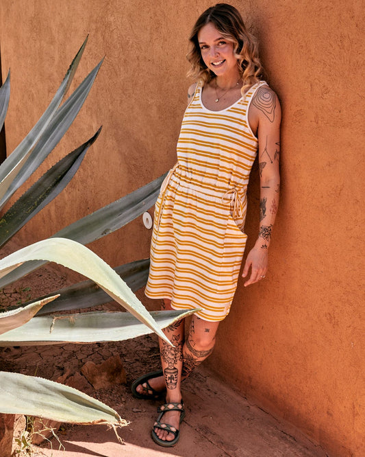 A woman in a knee-length, sleeveless Skylar Bauhaus - Womens Stripe Dress - Yellow smiles while standing by an orange wall, with a large agave plant in the foreground.