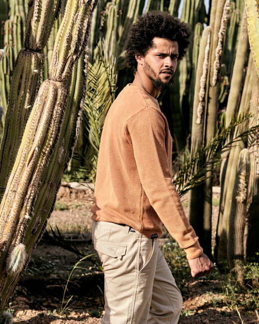 A man with curly hair walks past tall cacti, wearing a Saltrock Moss Mens Washed Knitted Crew in Orange and beige pants.