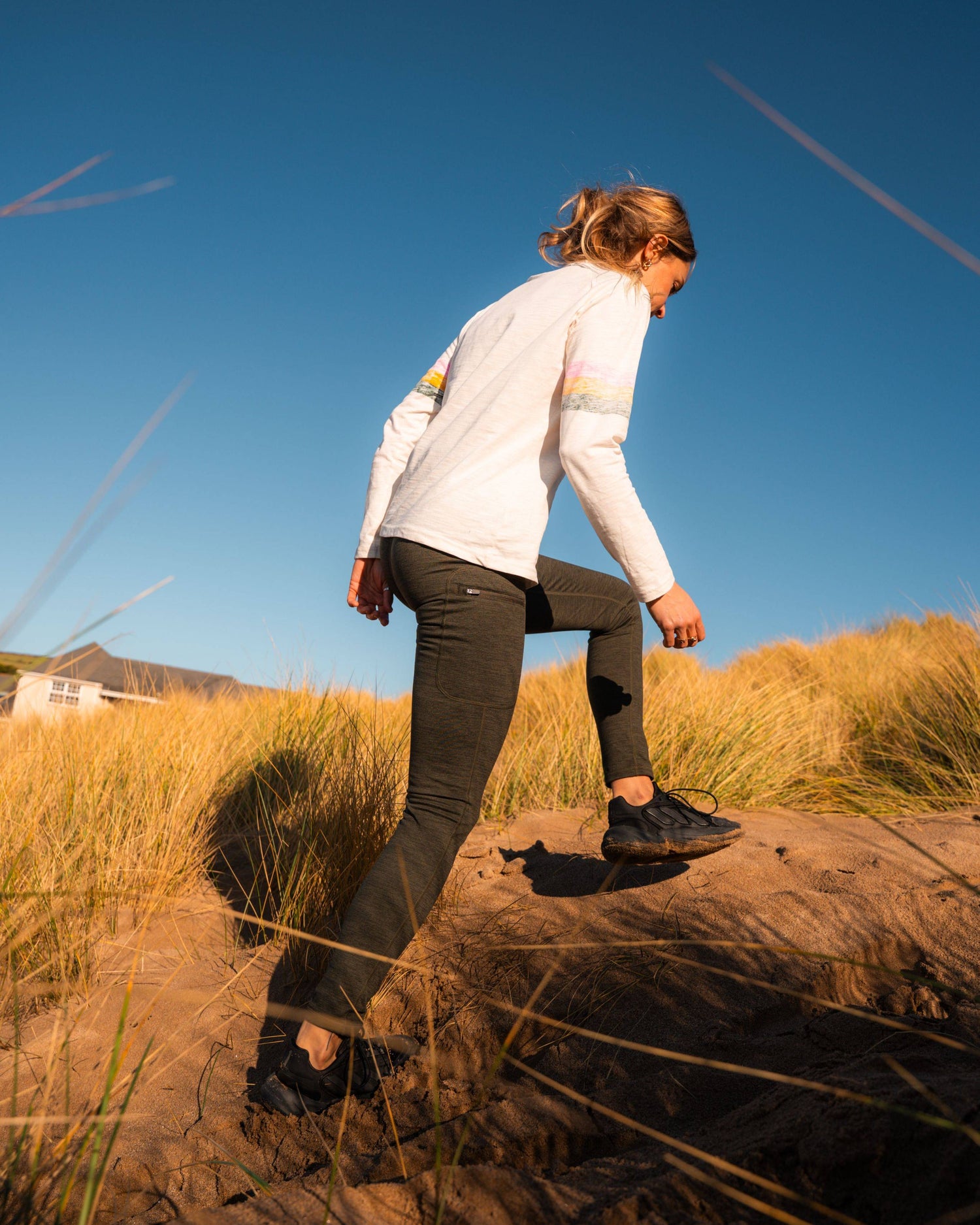 A woman in Saltrock Trek Womens 4 Way Stretch Outdoor Leggings in dark green walks uphill on sandy terrain, surrounded by tall grass and under a clear blue sky.