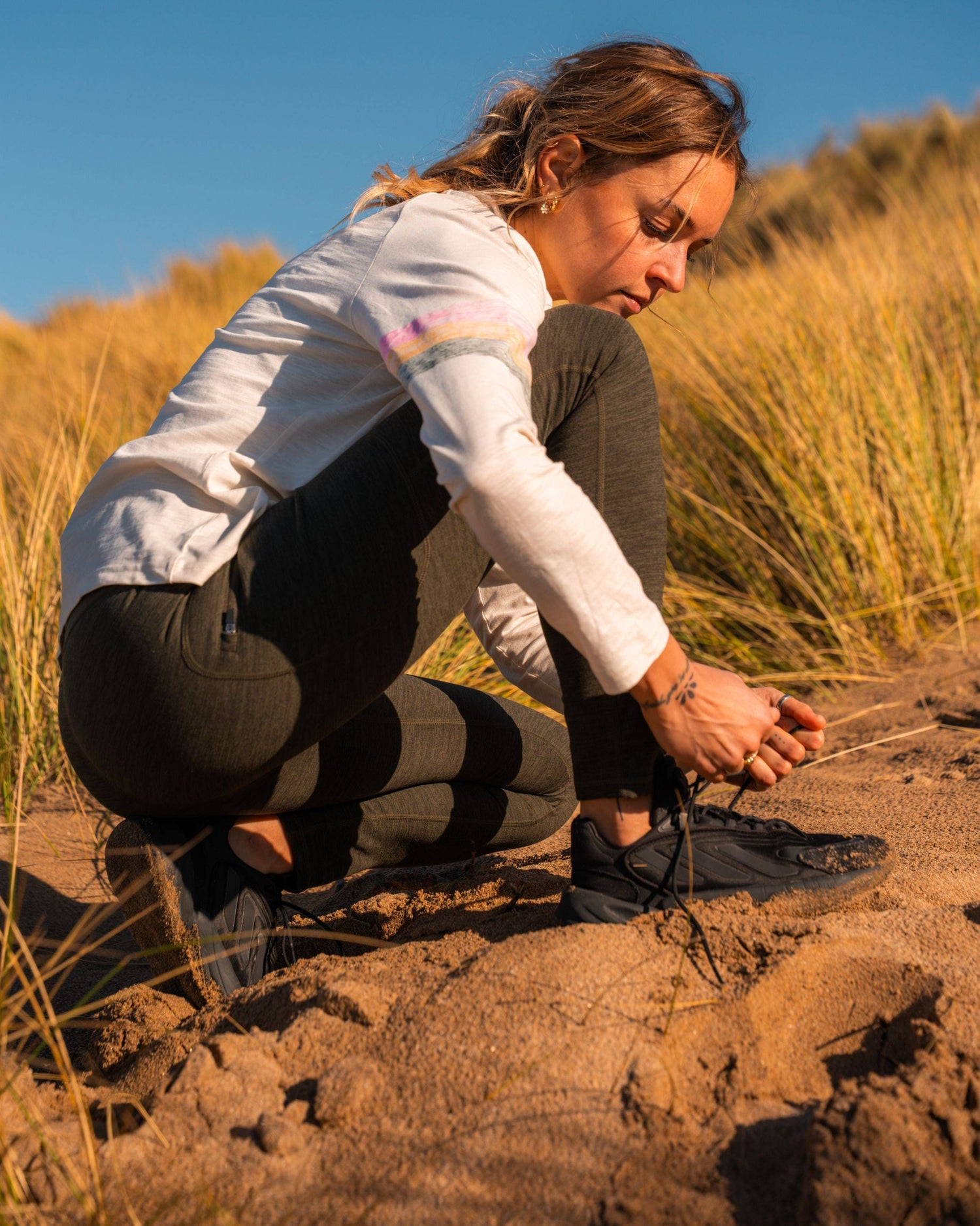 Wearing Saltrock's Trek Womens 4 Way Stretch Outdoor Leggings in dark green, a person in outdoor clothing crouches on sandy ground, tying black shoes. Their leggings have a side thigh zipped pocket, with tall grass and blue sky behind them.