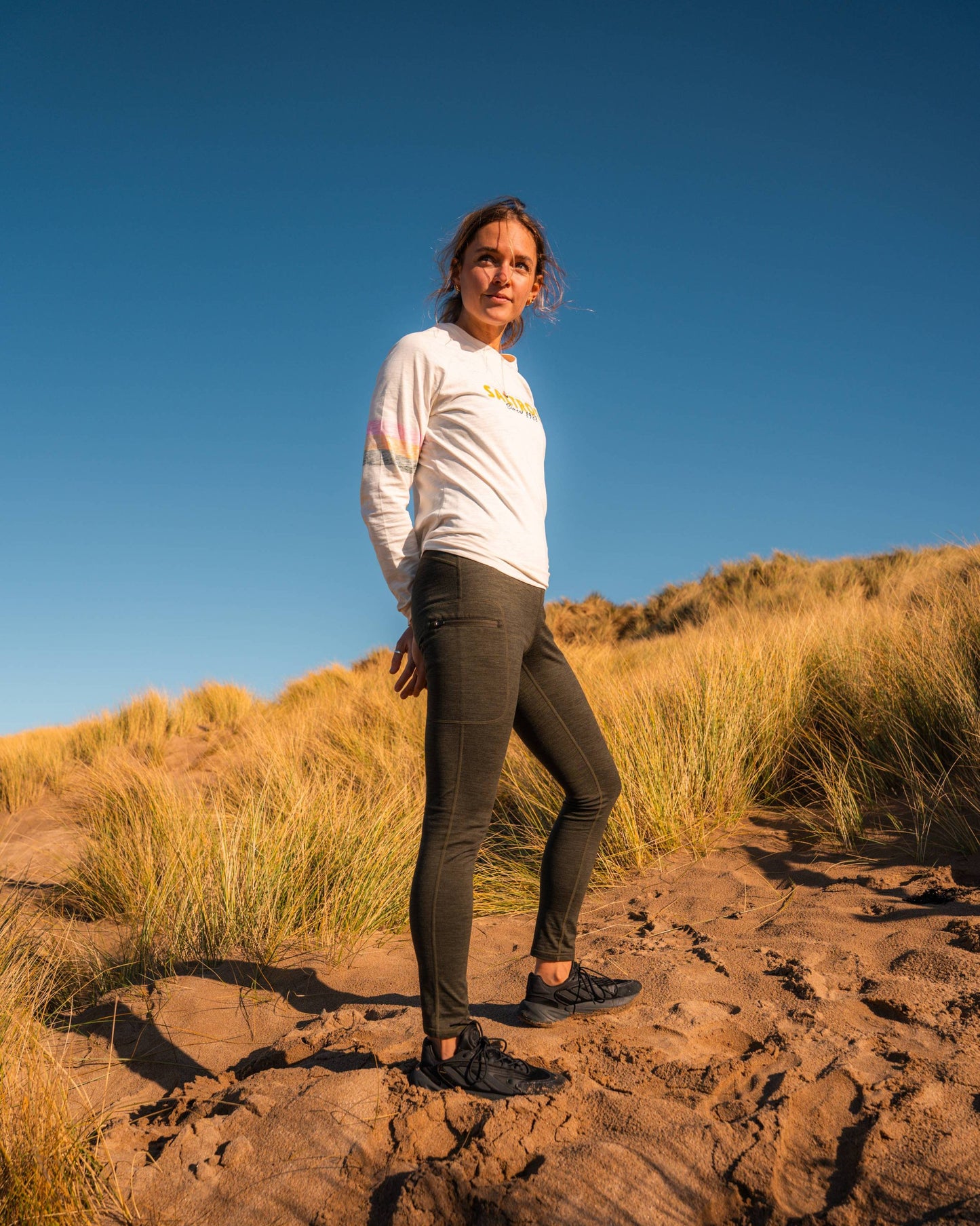 A person stands on sandy ground amid tall grass under a blue sky, wearing Saltrock Trek Women's 4 Way Stretch Outdoor Leggings in dark green, a white long-sleeve shirt, and black shoes.