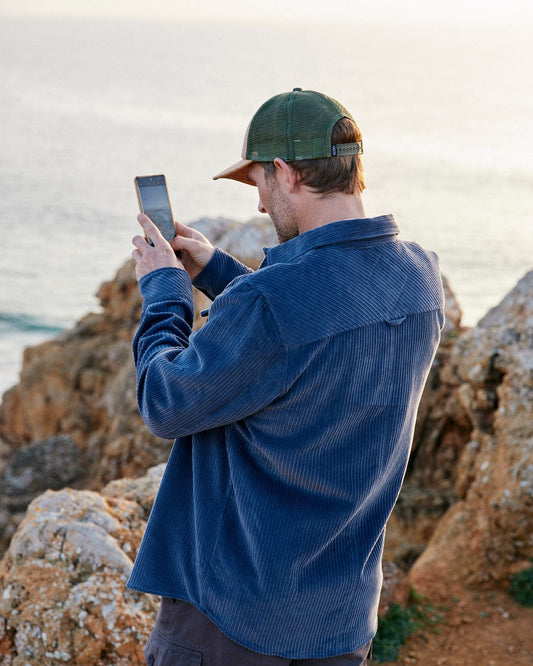 A man, wearing a Saltrock corduroy shirt with front chest pockets, is taking a picture of the ocean with his cell phone.