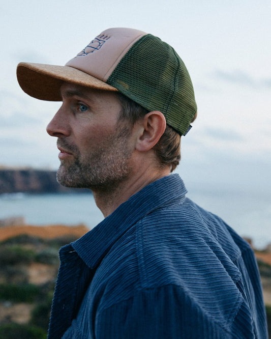 A man with stubble wearing a Saltrock Lineal Trucker Cap in Cream, boasting an adjustable strap and a mesh back, gazes into the distance with a serene backdrop of cliffs and sky.