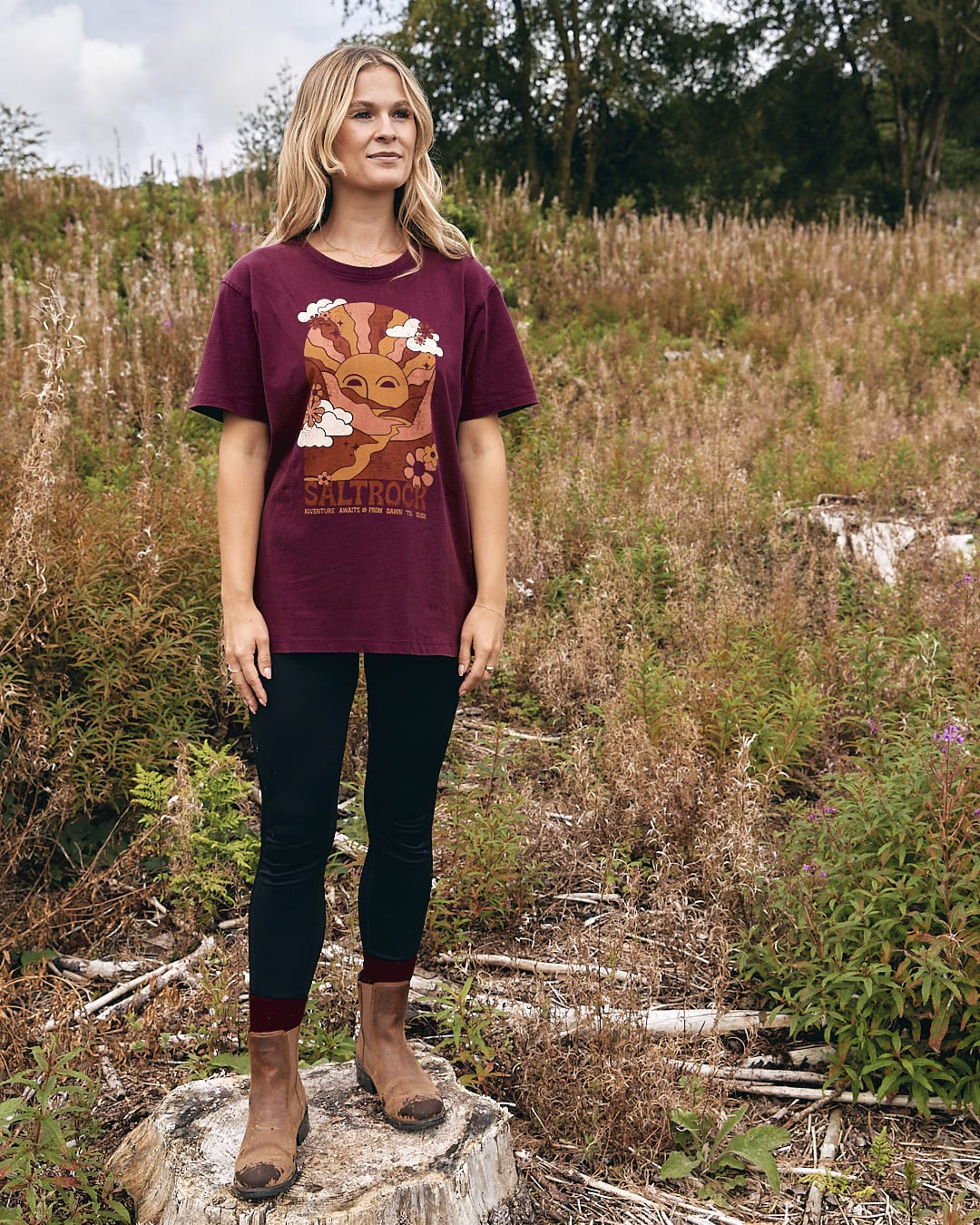 A woman wearing a Saltrock Adventure Awaits - Womens Short Sleeve T-Shirt - Dark Purple standing in a field.