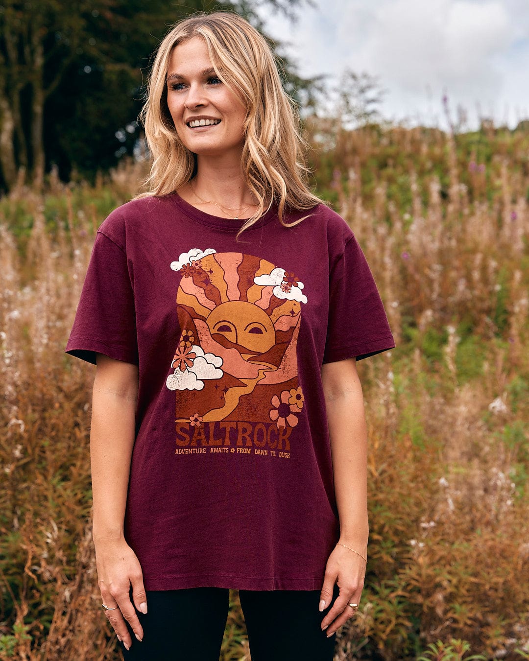 A woman wearing a Saltrock Adventure Awaits - Womens Short Sleeve T-Shirt - Dark Purple standing in a field.