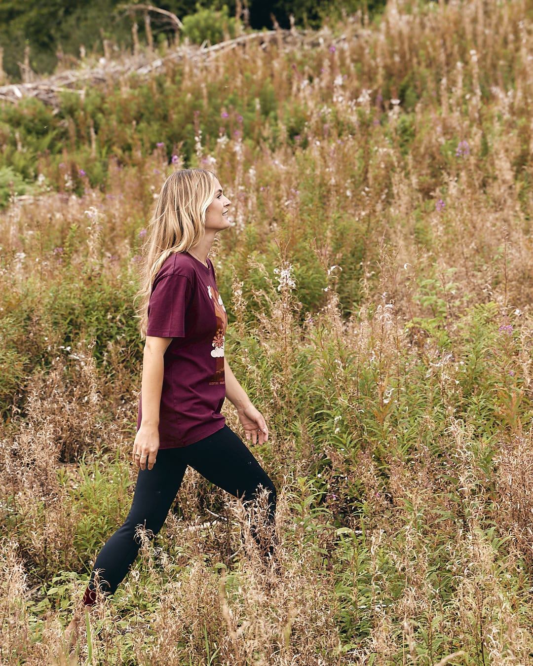 A woman walking through a field with an Adventure Awaits - Womens Short Sleeve T-Shirt - Dark Purple by Saltrock.