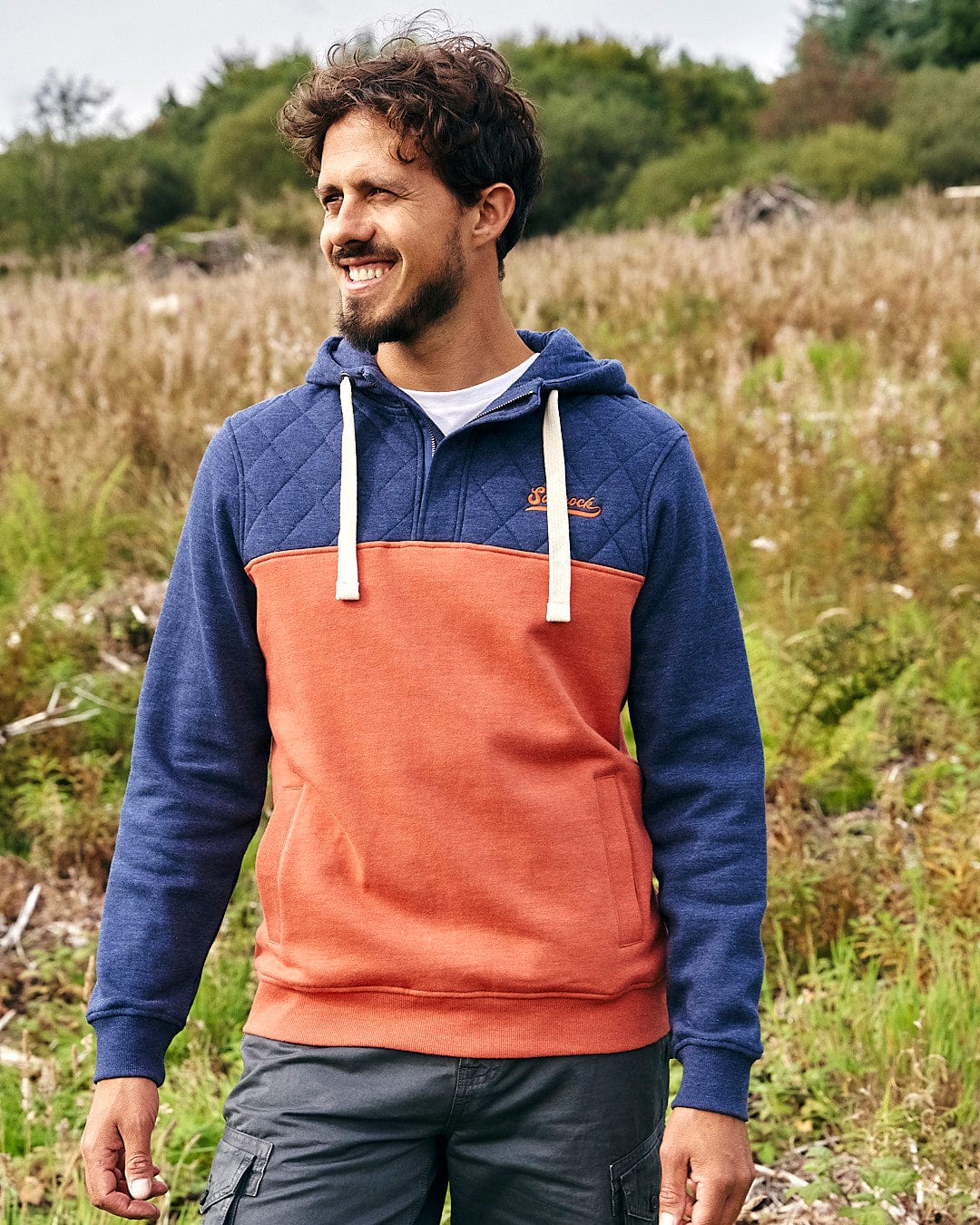 A man is standing in a field wearing a Saltrock - Aiken Mens 1/4 Neck Hoodie - Orange.