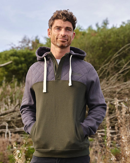 A man is standing in a field wearing the Saltrock Aiken Mens 1/4 Neck Hoodie - Green, enjoying warmth and comfort outdoors.