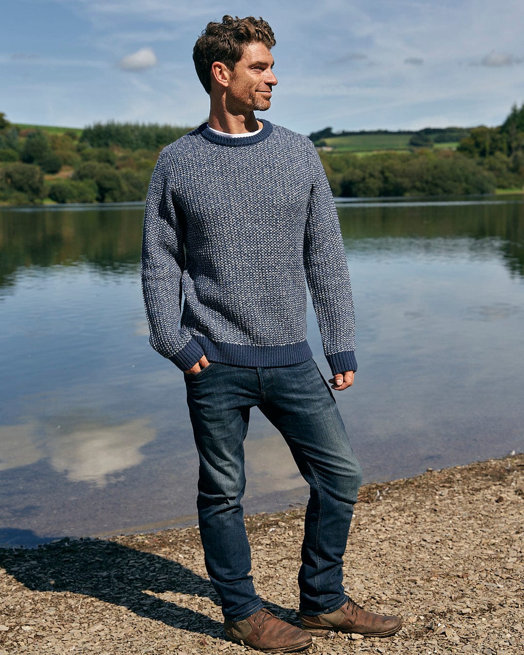 A man standing next to a body of water wearing a Saltrock Arlen - Mens Crew Knit - Dark Blue sweater and jeans.