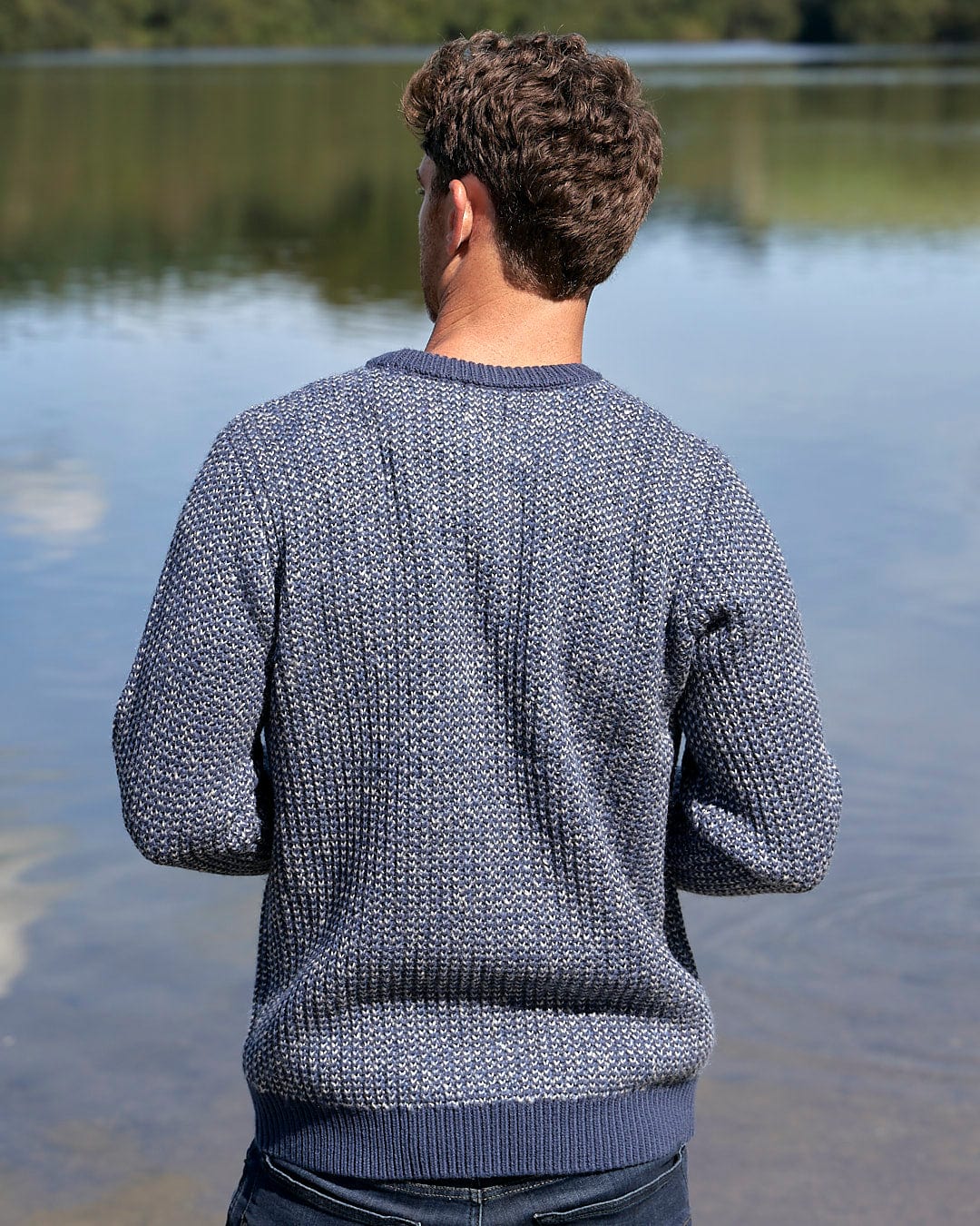 In cold weather conditions, a man is standing by a body of water wearing the Saltrock Arlen Mens Crew Knit Sweater in blue.