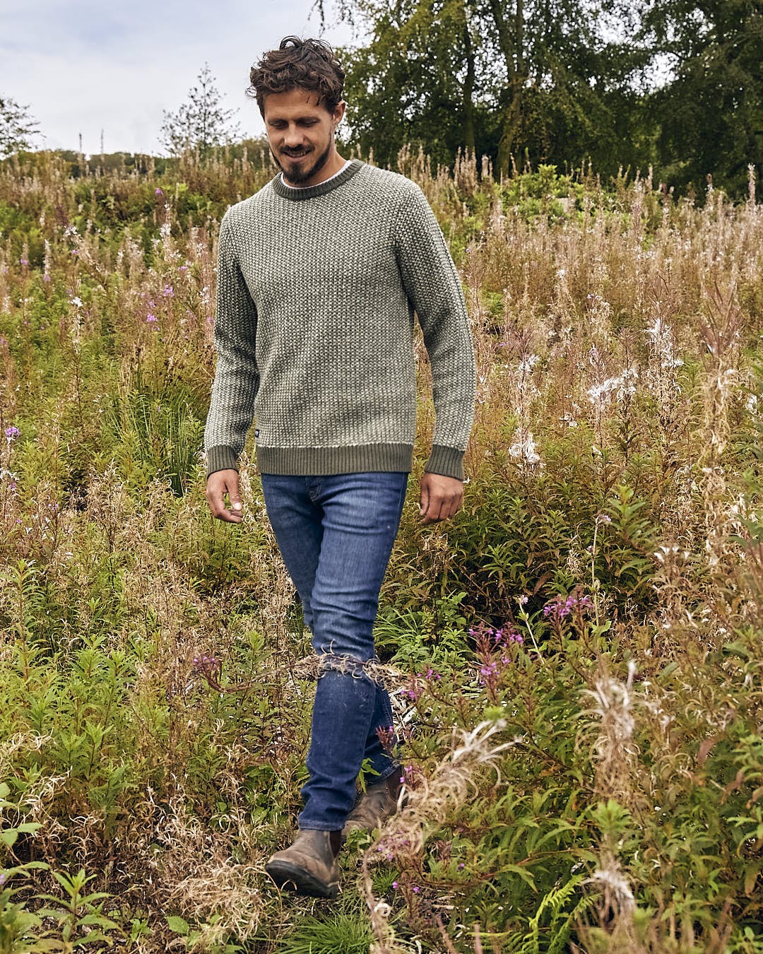 A man walking through a field of tall grasses wearing the Saltrock Arlen - Mens Crew Knit - Dark Green.