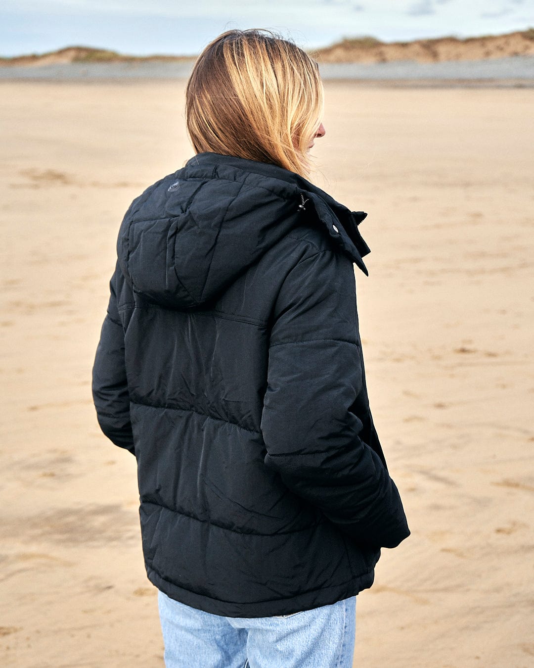 A woman standing on the beach wearing a Saltrock Aspen - Womens Short Puffer Jacket - Black.