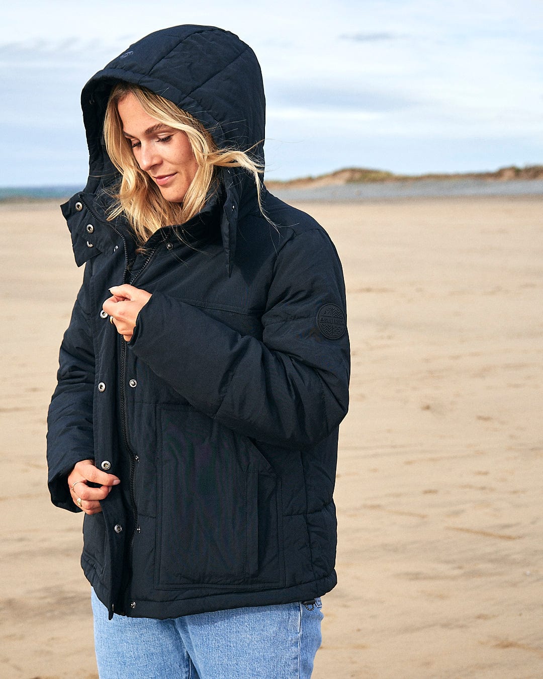 A woman wearing a Saltrock Aspen - Womens Short Puffer Jacket - Black on the beach.