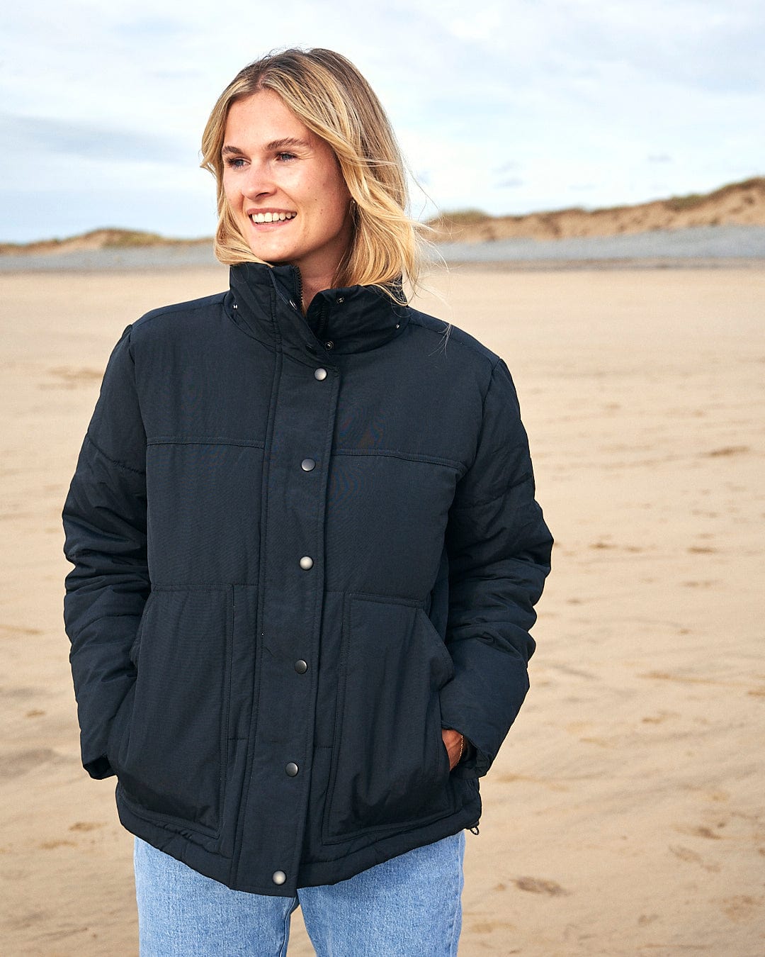 A woman is standing on a beach wearing a Saltrock Aspen - Womens Short Puffer Jacket - Black with a detachable hood.