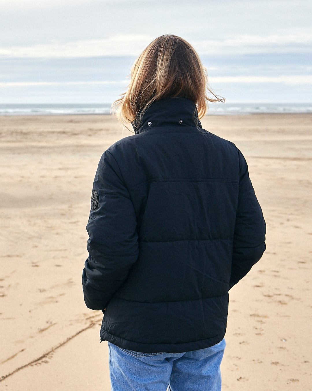 A woman standing on the beach wearing a Saltrock Aspen - Womens Short Puffer Jacket - Black with a detachable hood.