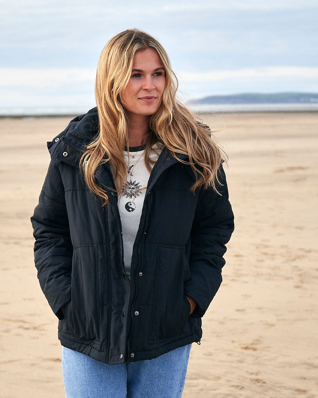 A woman standing on a beach wearing a Saltrock Aspen - Womens Short Puffer Jacket - Black with detachable hood.