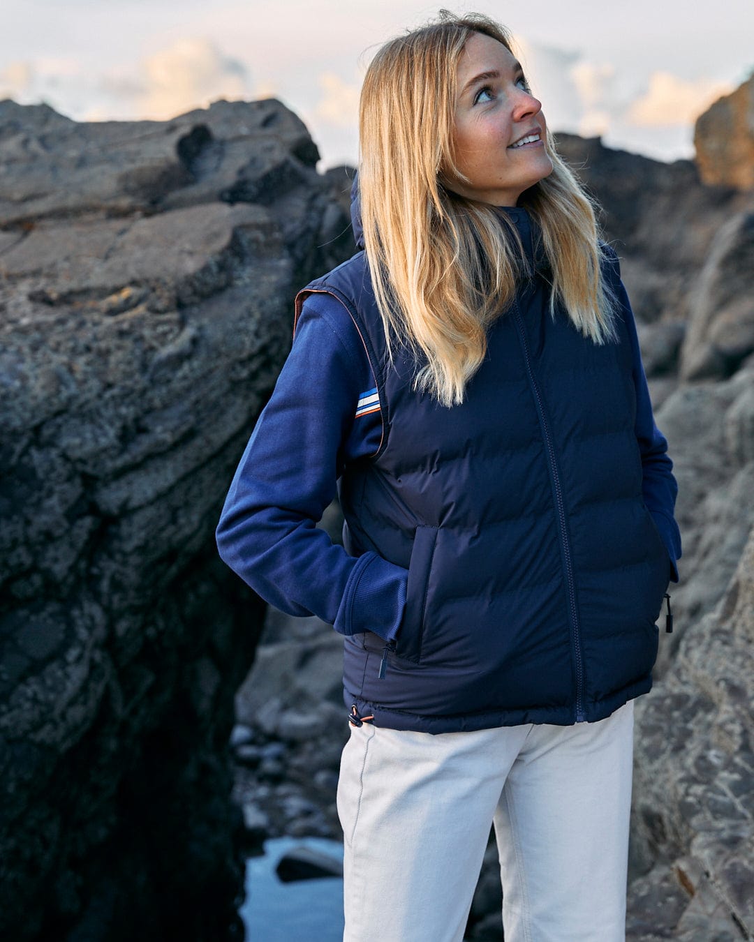 A woman in a Saltrock Astra - Womens Reversible Padded Gilet - Blue/Pink standing on rocks.