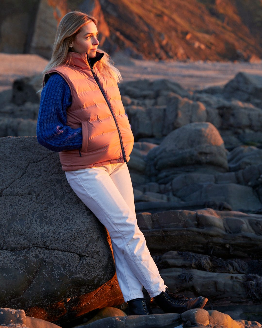 A woman is sitting on a rock, wearing the Saltrock Astra - Womens Reversible Padded Gilet - Blue/Pink.