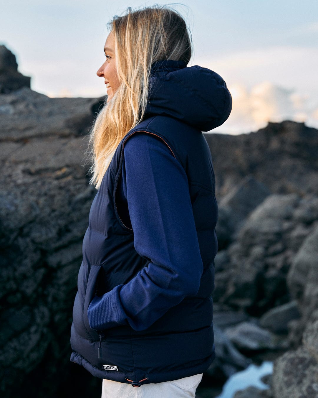 A woman wearing a blue puffer vest, the Saltrock Astra - Womens Reversible Padded Gilet - Blue/Pink, standing on rocks.