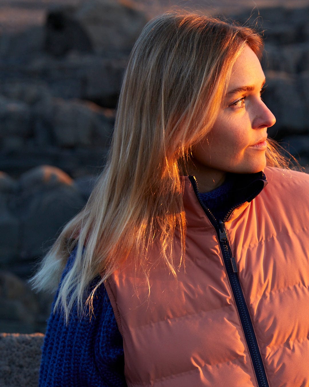 A woman wearing a Saltrock Astra - Womens Reversible Padded Gilet - Blue/Pink with a detachable hood on a rocky beach.