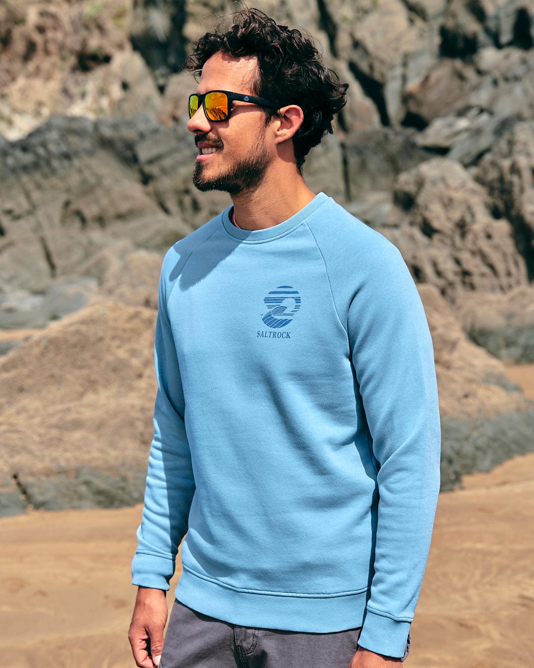 A man wearing sunglasses and a Saltrock Atlantic - Mens Crew Sweat - Light Blue standing on a beach.