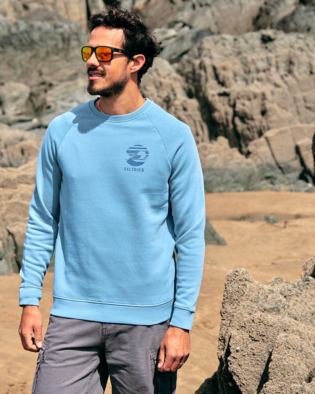 A man in a Saltrock Atlantic - Mens Crew Sweat - Light Blue sweatshirt standing on a rocky beach.