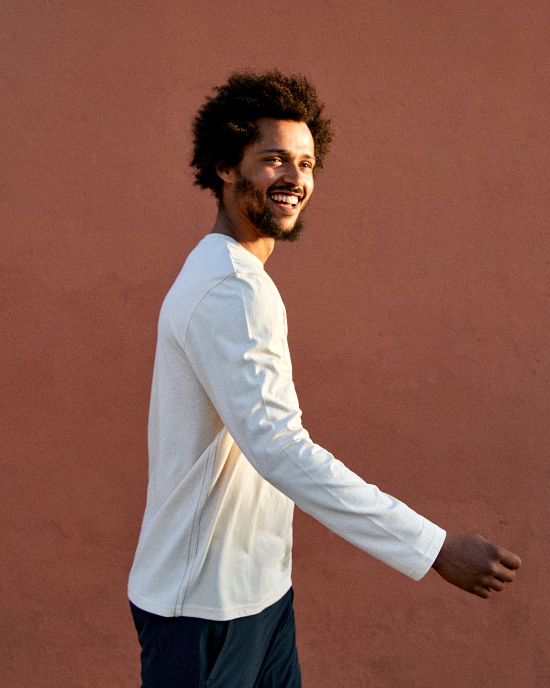 A young man with an afro smiling and walking past a pink wall, wearing a Saltrock Attendant Mens Long Sleeve T-Shirt in Cream and dark pants.