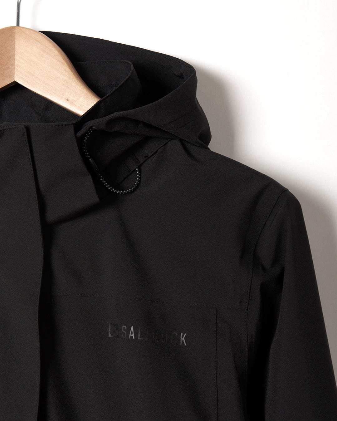 Close-up of a Saltrock Aubrey - Womens Waterproof Jacket - Black on a wooden hanger, displaying the brand logo "Saltrock" subtly embossed on the fabric.