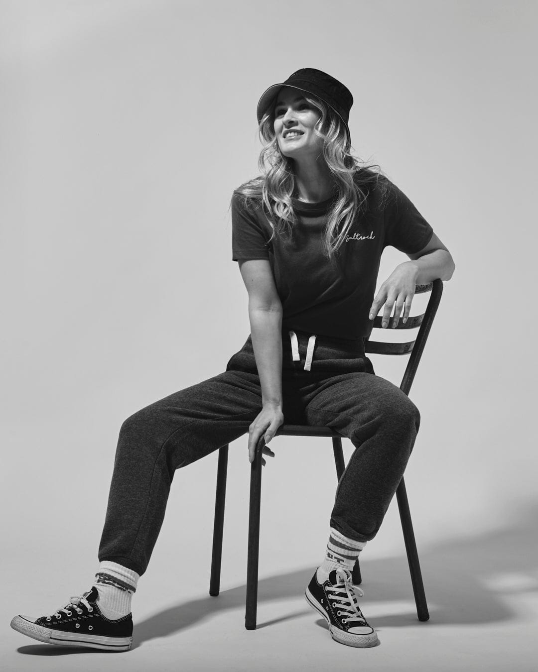 A woman in a casual outfit featuring Saltrock branding, with a Velator - Womens Joggers - Blue cap, sitting on a chair and smiling, in a monochrome studio setting.