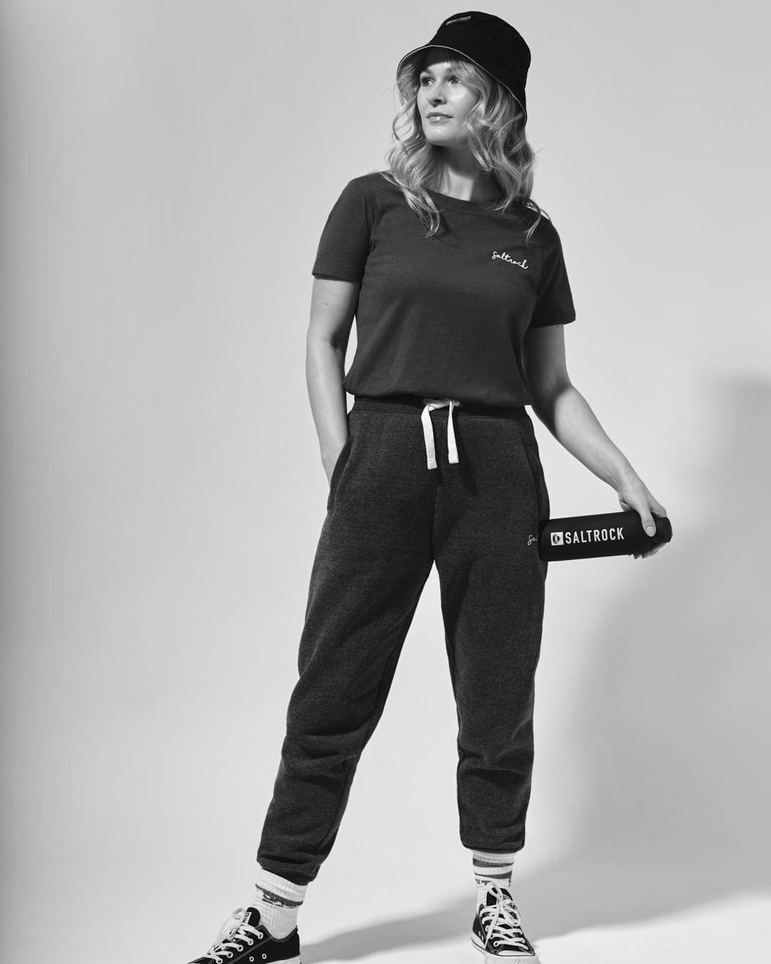 A woman in casual sportswear, including a cap and Velator Joggers in Blue with cuffed ankles, stands confidently in a black-and-white photo. She holds a book with "Saltrock" visible on the cover.