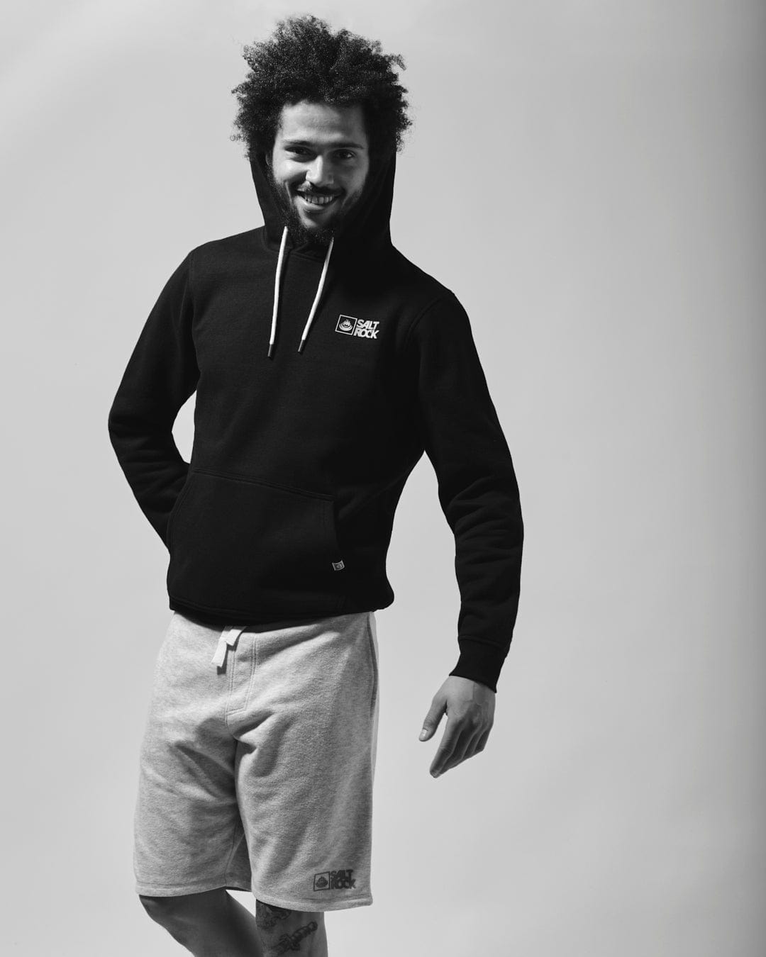 A person with curly hair wearing a Saltrock Original - Mens Pop Hoodie in Black made from soft jersey material and gray shorts stands with one hand in a pocket, smiling slightly at the camera.