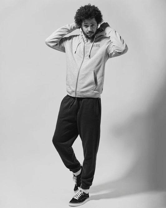 A young man with curly hair standing in a casual pose, wearing a grey hoodie with peach fluff lining, Saltrock Original - Mens Joggers - Black with an elasticated waist, and sneakers, against a plain background.