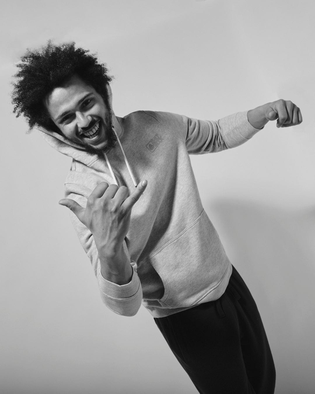 A man with an afro hairstyle joyfully posing in a Saltrock Original - Mens Pop Hoodie - Grey, making a gesture as if he's pretending to play guitar.
