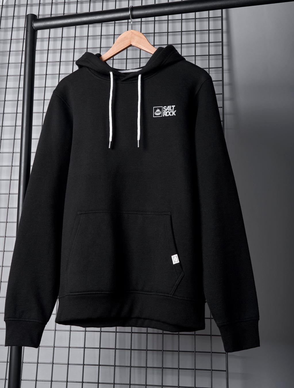 A Saltrock Original - Mens Pop Hoodie in Black, crafted from soft jersey material, with white drawstrings is displayed on a wooden hanger against a tiled wall background, featuring a small Saltrock branding logo on the left chest.