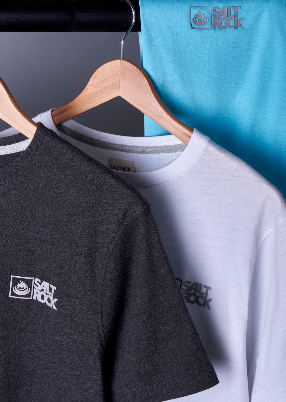 Three Saltrock Original - Mens Short Sleeve T-Shirts on hangers against a blue backdrop, featuring one black, one gray, and one blue shirt, each with the Saltrock branding.