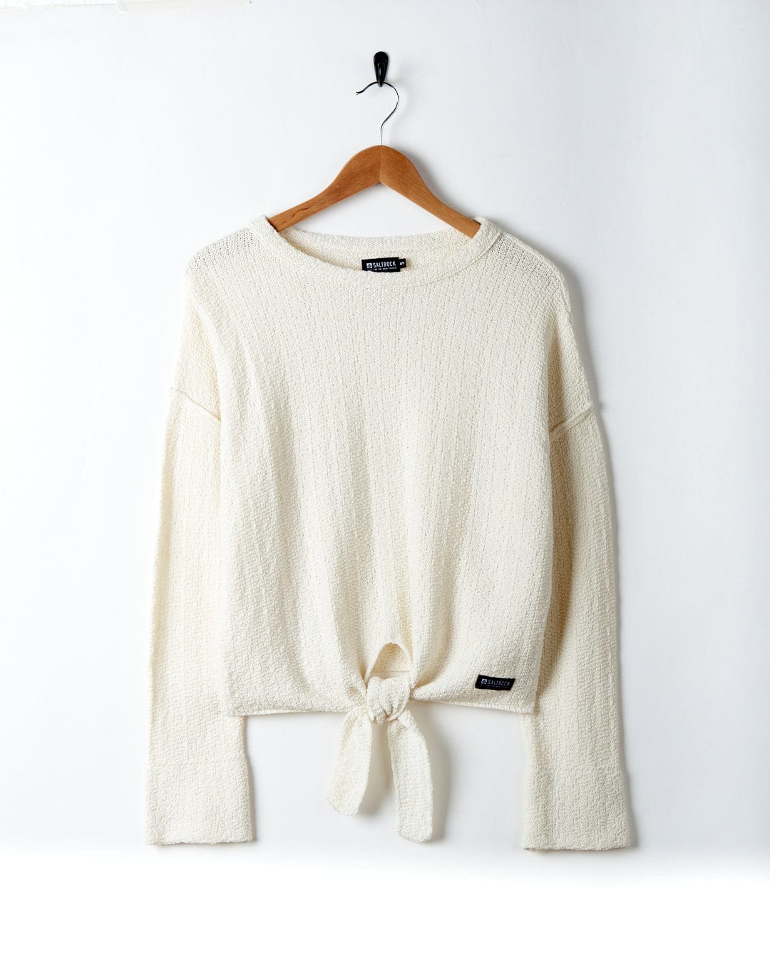 A Beachcomber - Womens Tie Waist Jumper - Cream by Saltrock, with a tie-front detail, hanging on a wooden hanger against a plain white wall.