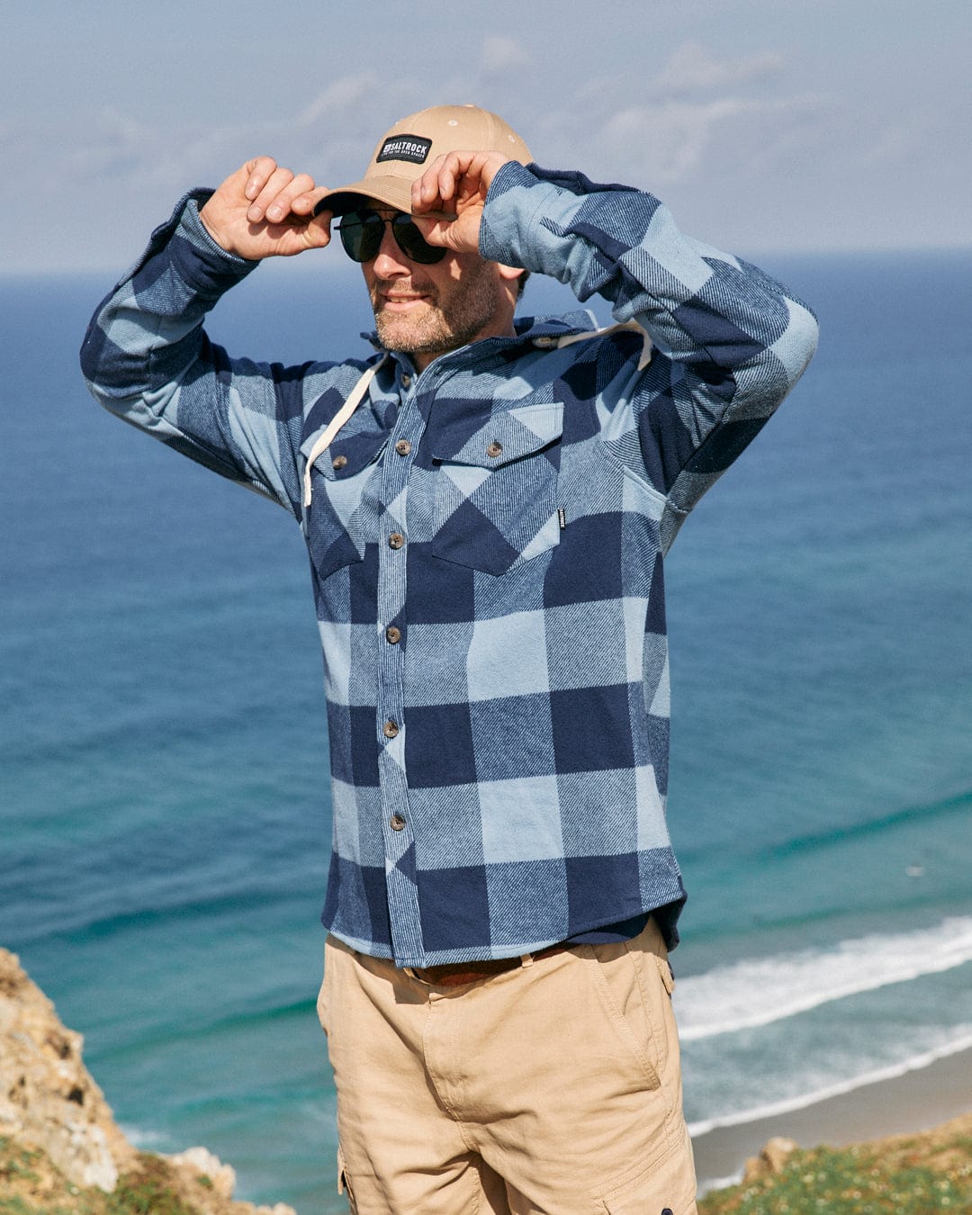 Man in a Saltrock Beale - Mens Hooded Long Sleeve Shirt - Blue and cap, looking towards the sea from a cliff, holding his sunglasses.