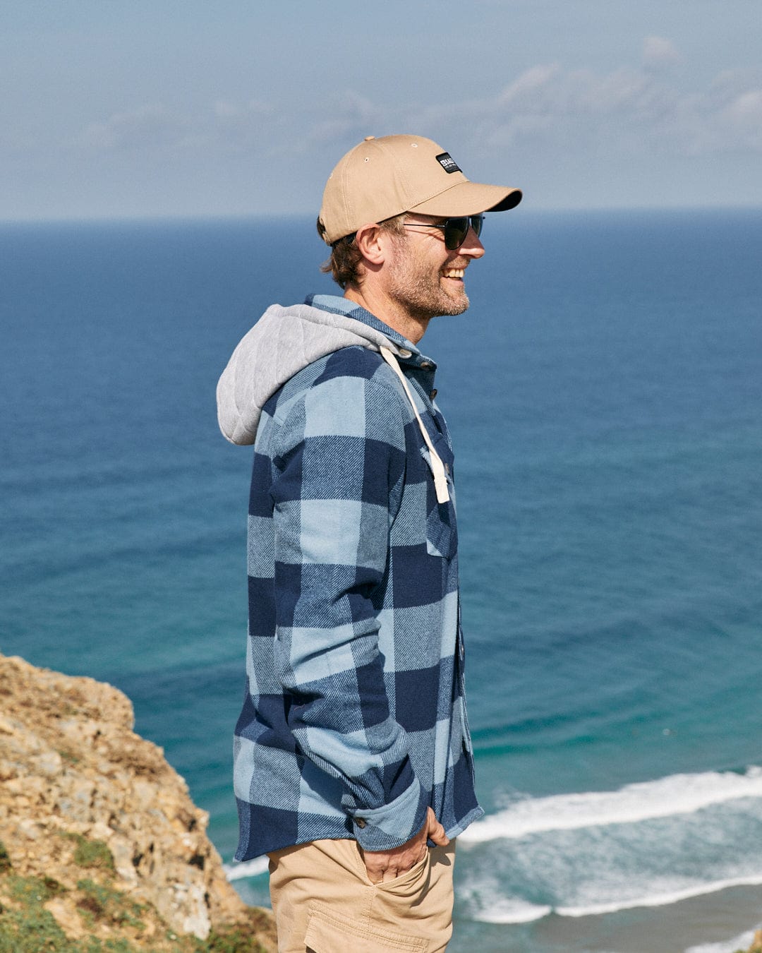 Man in a Saltrock Beale - Mens Hooded Long Sleeve Shirt - Blue and cap smiling while looking out over the sea from a coastal cliff.