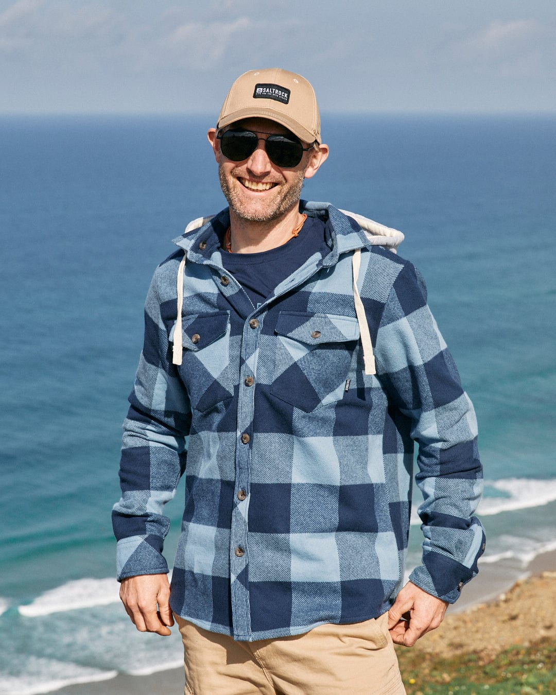 Man in sunglasses and a cap, wearing a Saltrock Beale - Mens Hooded Long Sleeve Shirt in Blue with a detachable hood, smiling by the seaside.