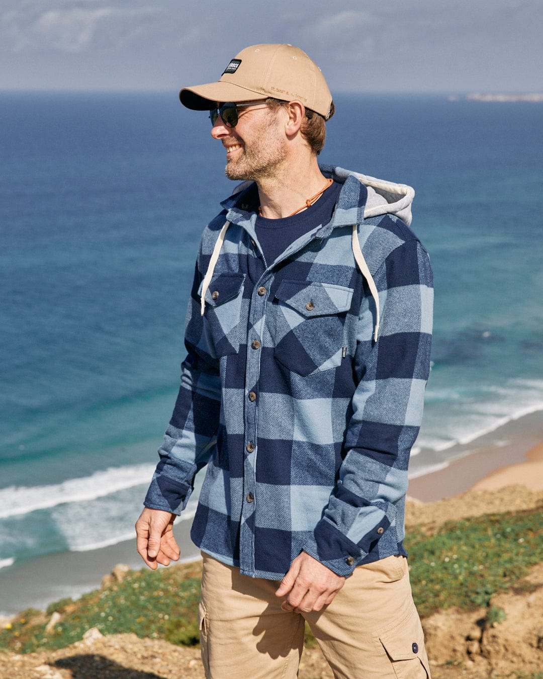 Man wearing a cap, Saltrock Beale - Mens Hooded Long Sleeve Shirt in Blue with a detachable hood, and sunglasses smiling while looking to the side, with a beach and the ocean in the background.