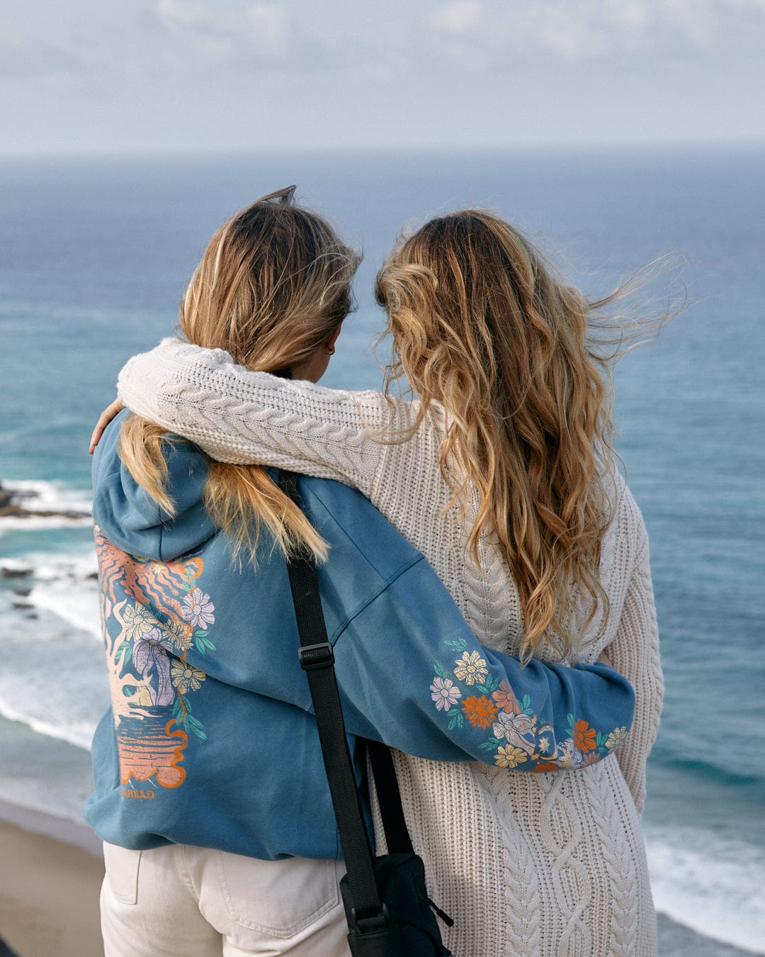 Two women are standing close to each other, overlooking a sea view, with Luna's arm around the other's shoulder. Both are wearing Saltrock sweaters and have wavy blonde hair.