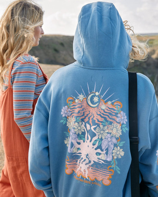 Two people outdoors, one facing away from the camera wearing a Saltrock blue "Better Days" Womens Pop Hoodie with an intricate floral design, and the other facing the camera, partially obscured, wearing a striped top and an orange jumpsuit.