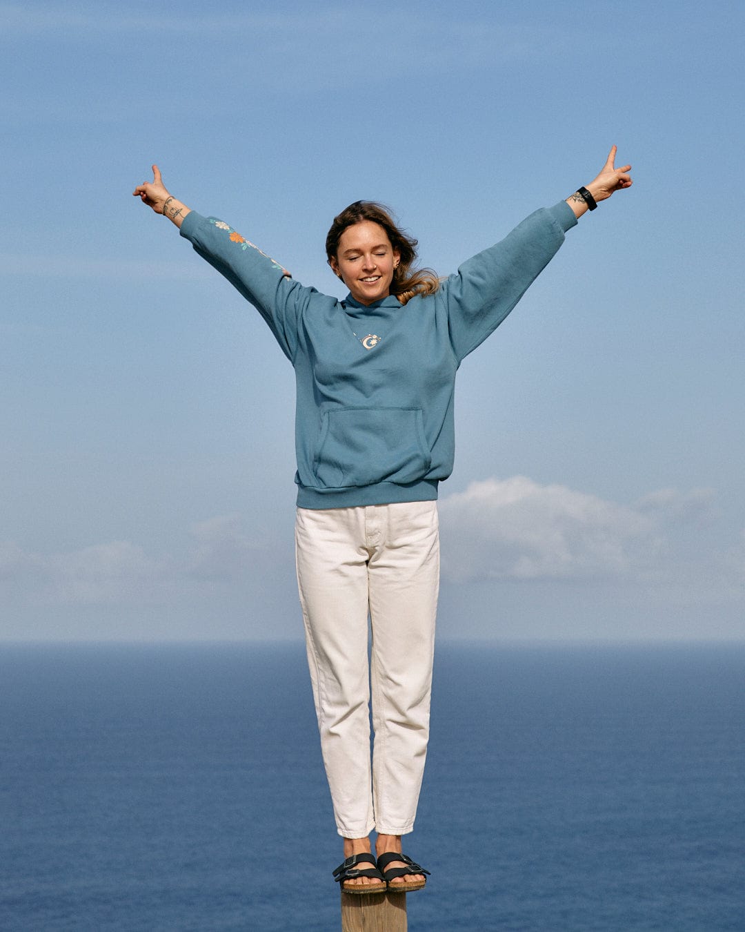 A person in a Better Days - Womens Pop Hoodie in blue and white pants stands on a ledge with their back to the sea, arms raised and making a victory sign with both hands, embodying the spirit of "Brighter Days" by Saltrock.