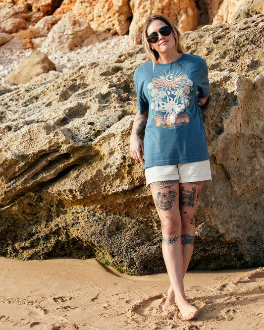 Woman standing on a sandy beach, leaning against a rocky cliff, wearing a Saltrock Better Days Recycled Womens Short Sleeve Relaxed T-Shirt in Teal and shorts made of recycled polyester, with visible leg tattoos.
