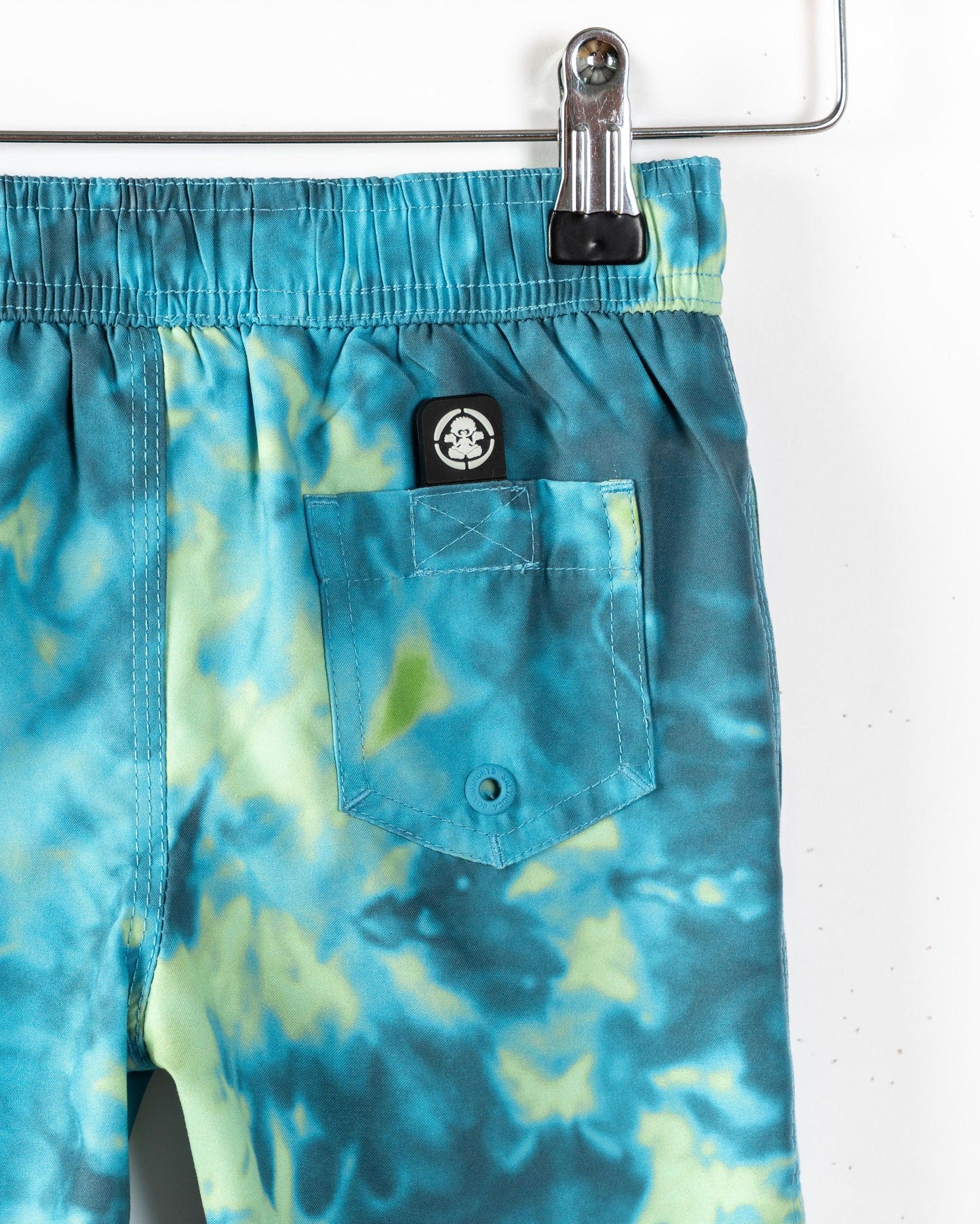 Close-up of Saltrock Ocean Swim Kids Swim Shorts in blue with a tie-dye print, elasticated waist, buttoned back pocket, and a black and white Saltrock logo above the pocket, hanging on a metal hanger.
