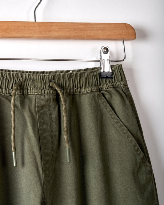 Close-up of Saltrock's Bora Bora Kids Chino Trouser in dark green, featuring a baggy fit and elastic waistband, displayed on a wooden hanger against a white background.