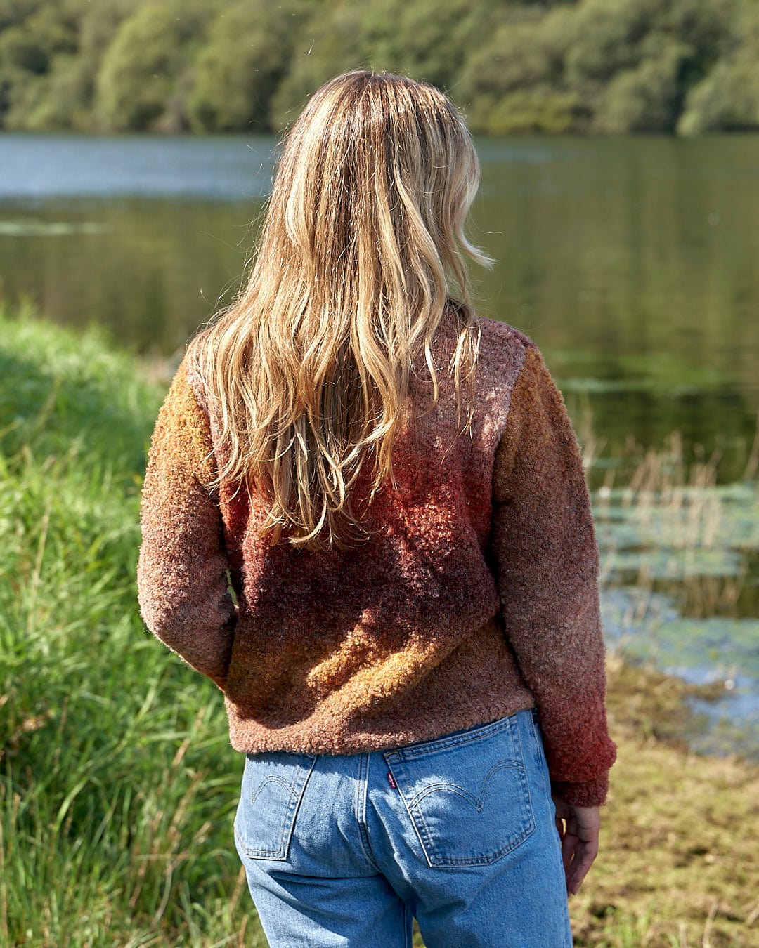 A woman wearing the Saltrock Bowden - Boucle Crew Jumper - Orange and jeans is standing by a lake.