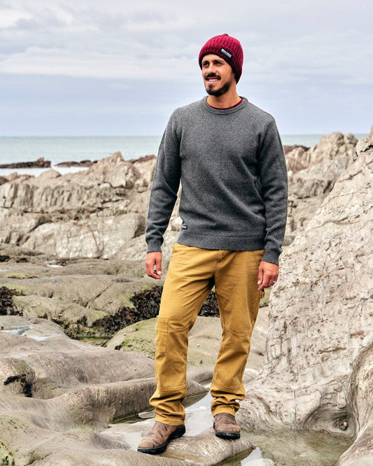 A man standing on rocks near the ocean wearing a beanie and khaki pants, showcasing the Saltrock branding of the Bowen - Mens Knitted Crew - Grey.